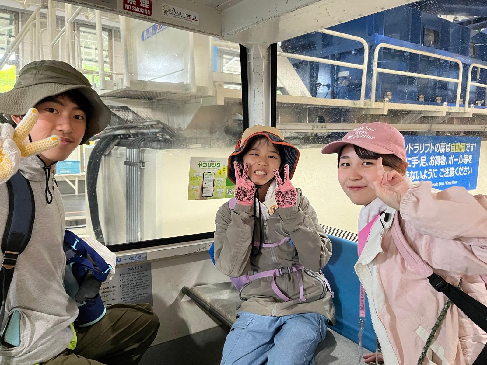 ゴンドラの車内。帽子をかぶった子どもとリーダーが座席に座り、カメラに向かっている。窓の外にはゴンドラ乗り場の鉄骨が見える