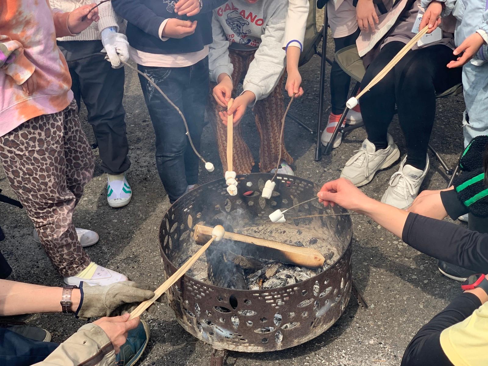 屋外の焚き火台を囲み、子どもたちとリーダーが枝や割り箸に刺したマシュマロを火にかざしている