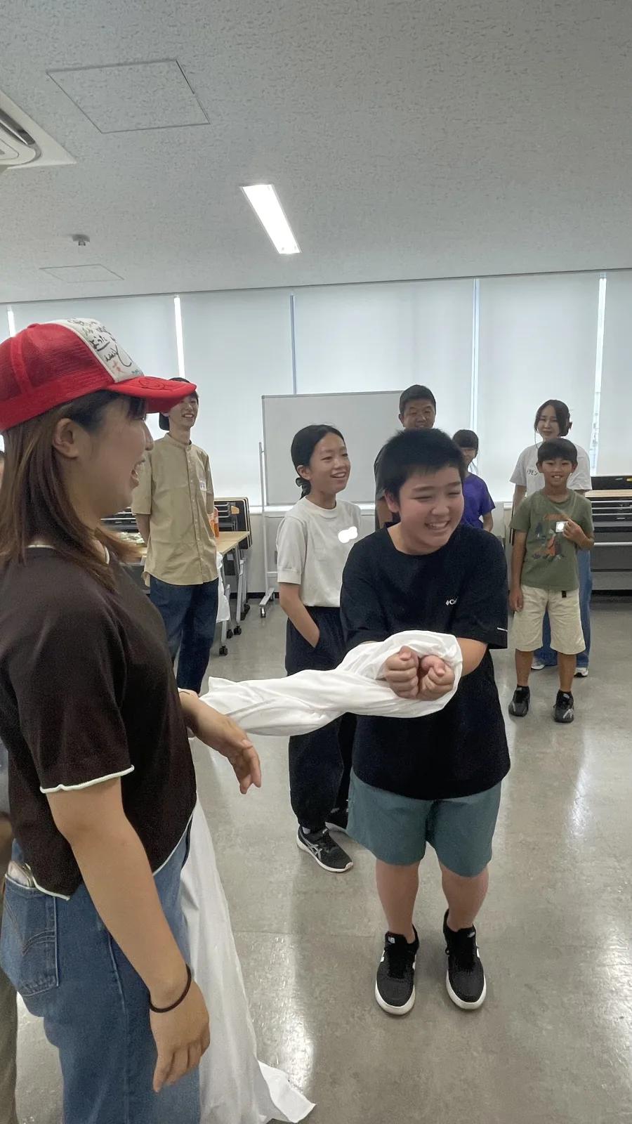 白い布を両手首に巻きつけて手錠の形にした子どもと、それを見て笑っている子どもたちとスタッフ