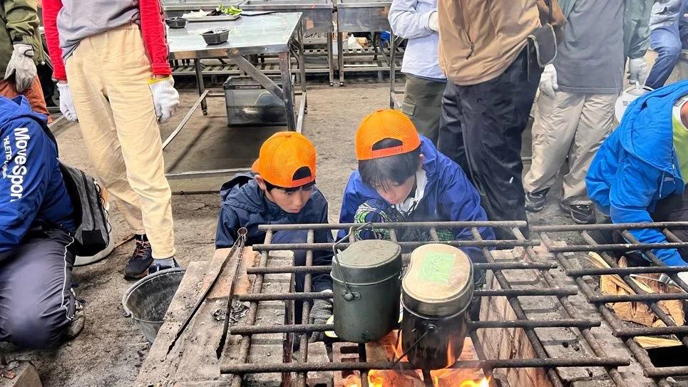 野外炊事場の焚き火の上に鉄の網が渡され、飯ごうが2つかけられている。オレンジのキャップをかぶった参加者ふたりが、しゃがんで火の様子をのぞき込んでいる
