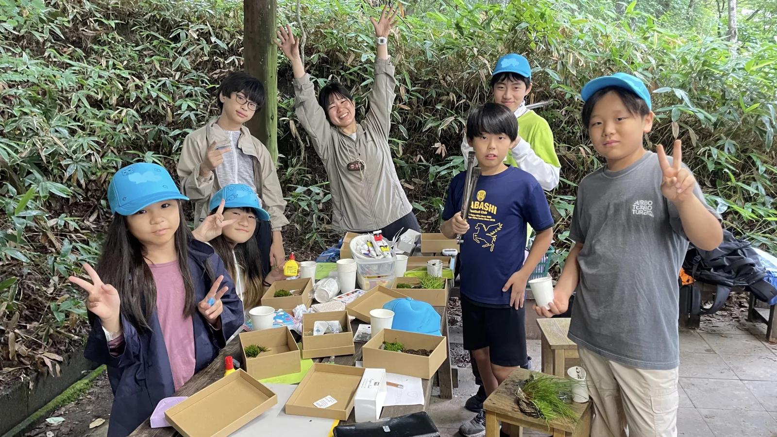 屋根付きの小屋のテーブルに、苔や土を敷いた紙箱がいくつも並んでいる。青いキャップの参加者とスタッフが、道具を手に囲んで笑っている