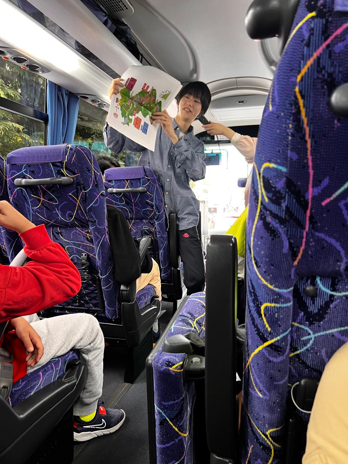 観光バスの車内。通路に立ったリーダーがマイクを持ち、山のイラストが描かれた紙を掲げて説明している。座席には子どもたちが座っている