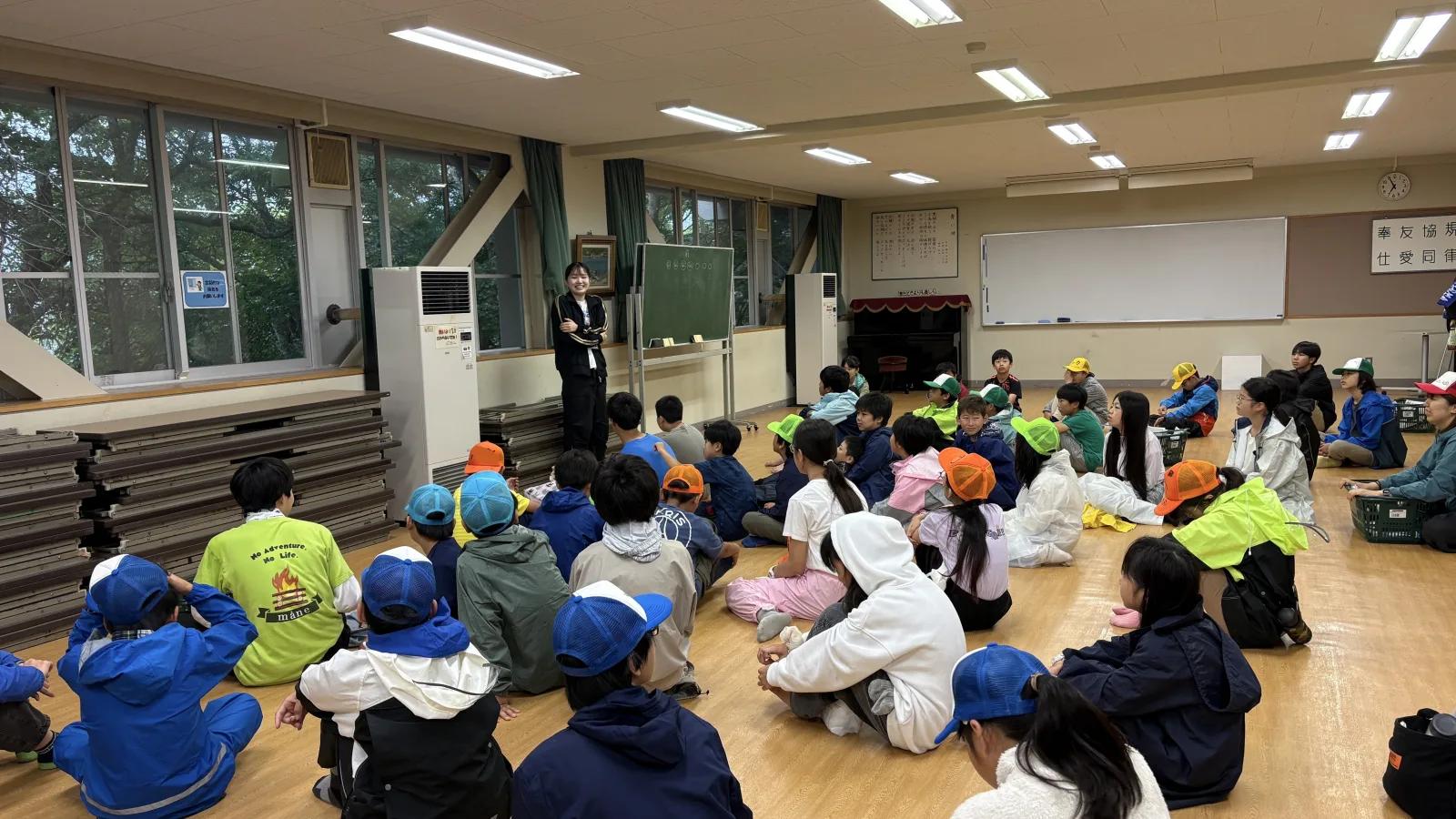 施設の広間。色とりどりのキャップをかぶった参加者が床に座り、前に立ったスタッフの話を聞いている。窓の外は緑