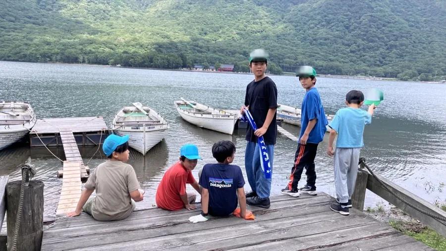 桟橋に腰かけたり立ったりしている参加者6人。手すりの向こうに手こぎボートが並び、対岸には山と朱塗りの建物が見える