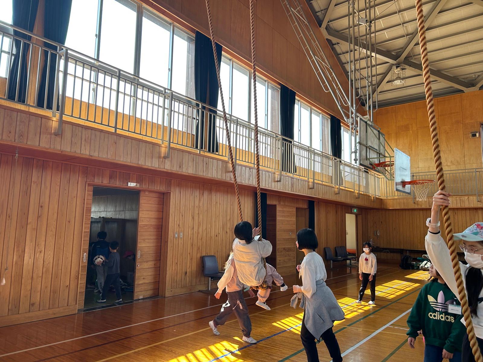 体育館の登り綱に、子どもがぶら下がって揺れている。午後の光が差し込んでいる