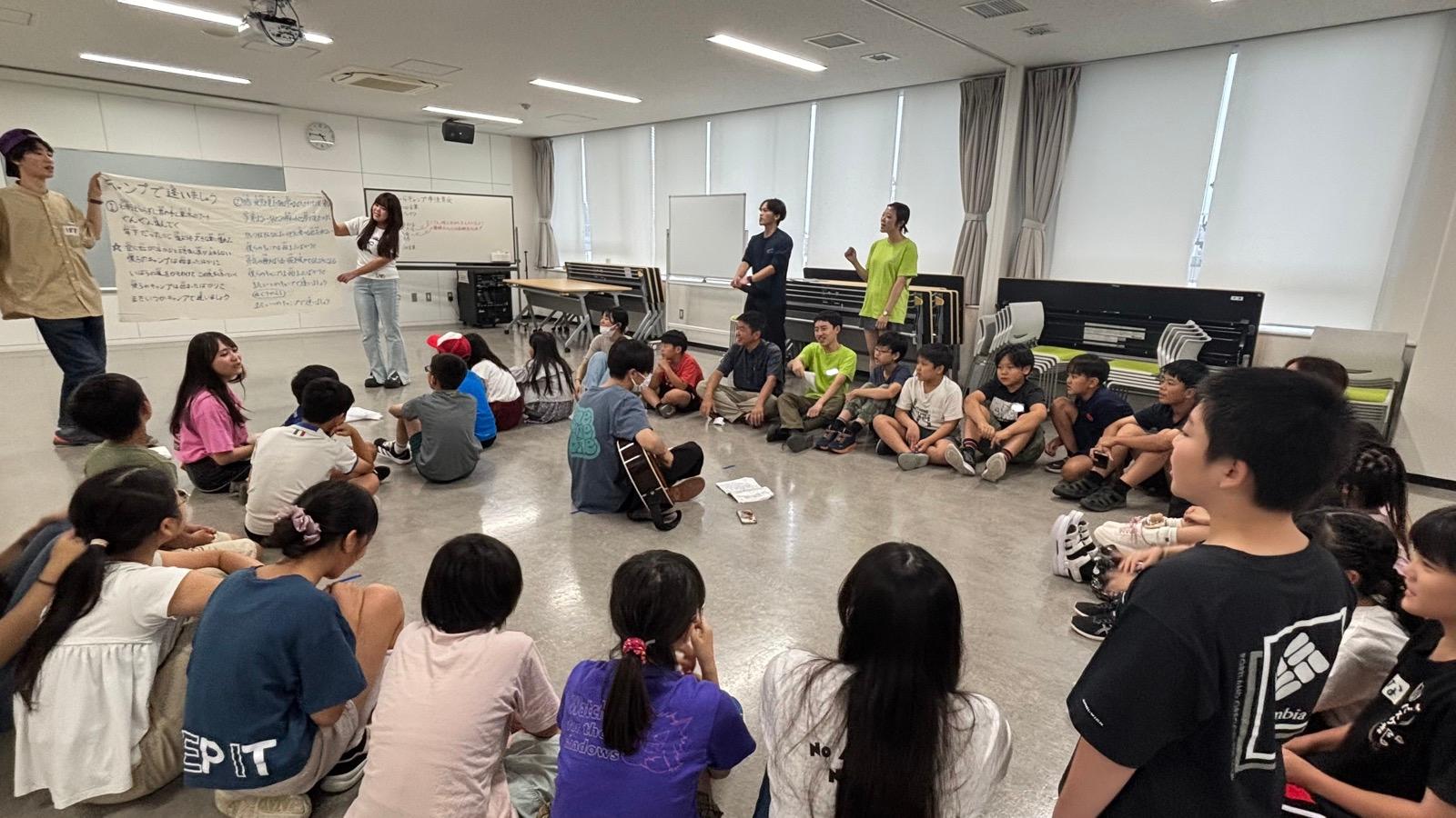 会議室で全員が輪になって床に座り、中央でギターが弾かれている。左側では歌詞を書いた大きな紙が広げられ、「キャンプで逢いましょう」と書かれている