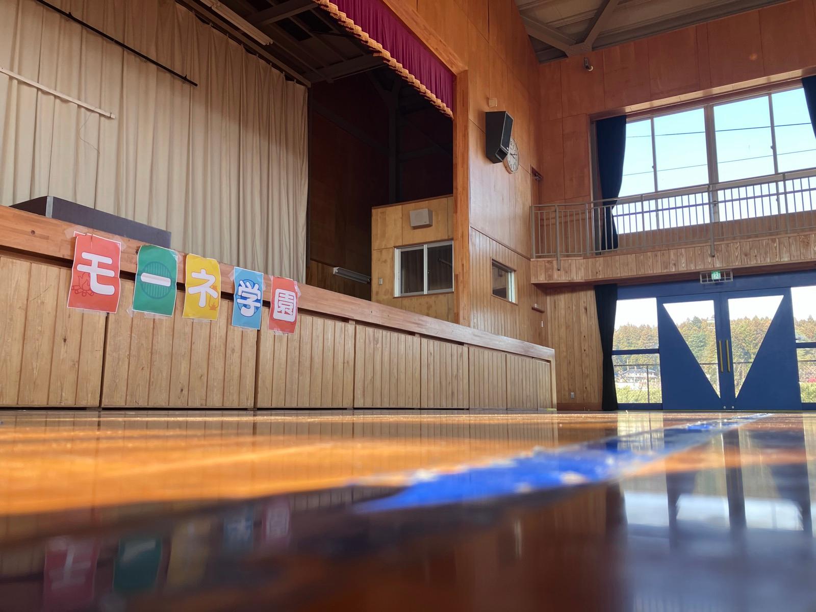 誰もいない体育館を床すれすれの低い位置から撮った写真。舞台の前に「モーネ学園」の文字が貼られている