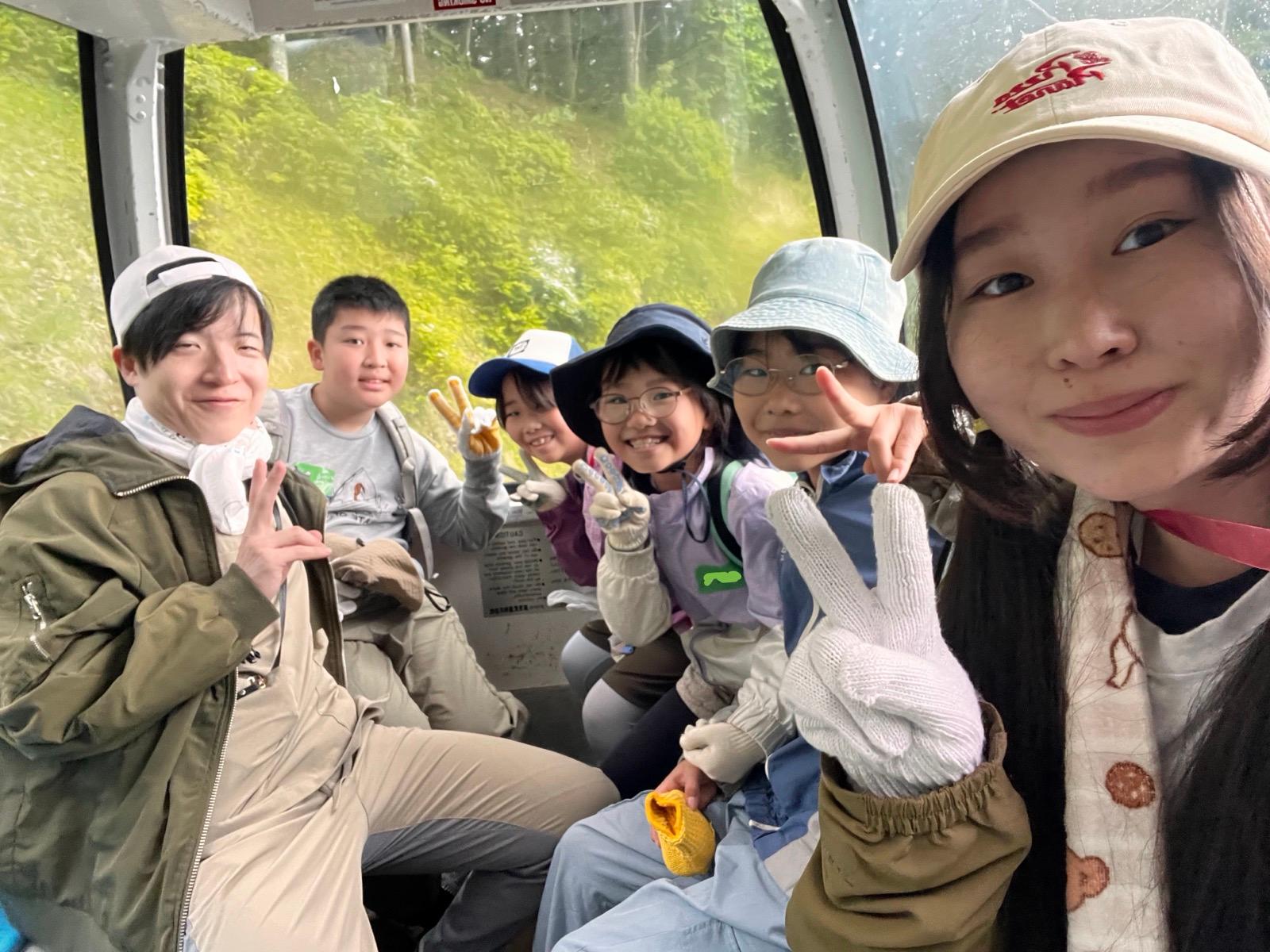 ゴンドラの車内。リーダーと子どもたちが並んで座り、カメラに向かってピースをしている。子どもは軍手をつけている