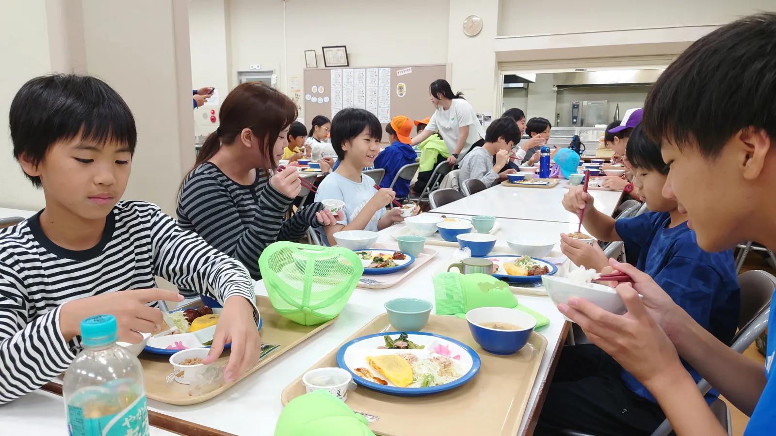 施設の食堂。長机に並んだトレーの朝食を食べる参加者たち。ごはん、汁物、おかずの小皿が乗っている