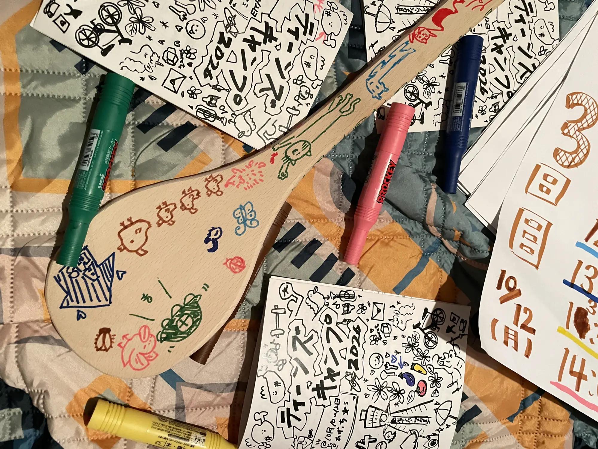 ティーンズキャンプのしおりとマーカーのそばに、参加者が絵を描いた木のしゃもじが置かれている様子